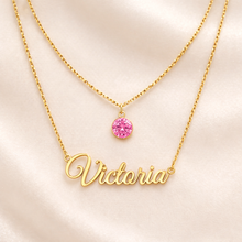 Load image into Gallery viewer, Gold necklace with 'Victoria' and pink gemstone on a light pink background