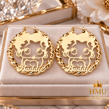 Load image into Gallery viewer, A pair of gold hoop earrings displayed on a cream velvet jewelry box. Each earring features a twisted rope border and a central cutout design of a cartoon character with the name 'Baddie' written in script.