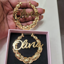 Load image into Gallery viewer, Gold hoop earrings with 'Olivia' engraving held above a pink box containing another pair of similar earrings.