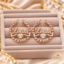 Load image into Gallery viewer, A pair of rose gold hoop earrings with twisted rope borders and the name 'Lolita' in script, displayed on a beige velvet jewelry box. The earrings feature a small cartoon character cutout.