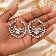 Load image into Gallery viewer, A close-up of a hand holding a pair of large silver hoop earrings personalized with the name 'Tiana' and a small Bettt Boop face. The background shows draped cream fabric and gold decorative elements, with an HMU LADIES logo in the bottom right corner.