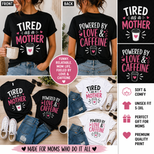 Load image into Gallery viewer, Black t-shirt with 'Tired as a Mother, Powered by Love &amp; Caffeine' text and graphics, worn by a person.
