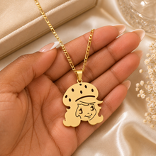 Load image into Gallery viewer, A gold pendant necklace featuring a cartoon girl with a hat, held in a hand over a beige satin fabric background with dried flowers.
