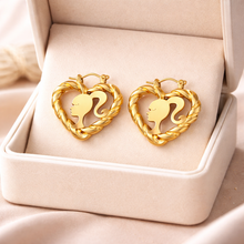 Load image into Gallery viewer, Gold heart-shaped earrings with a Barbie silhouette design on a beige jewelry box.
