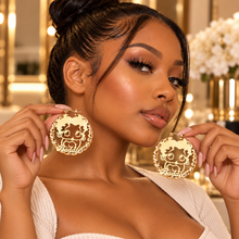 Load image into Gallery viewer, A woman holds up a pair of large gold-tone circular earrings featuring a lion silhouette and the word 'Baddie" in a raised font, shown against a warm, softly blurred indoor background.

