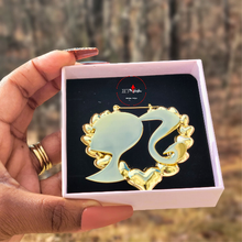 Load image into Gallery viewer, Gold Barbie head heart-shaped earring in a box held by a hand with brown nail polish.
